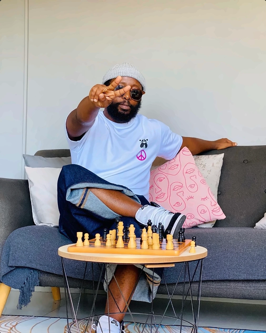 A man in a white SACXESS tee and grey beanie making a peace sign beside a chessboard