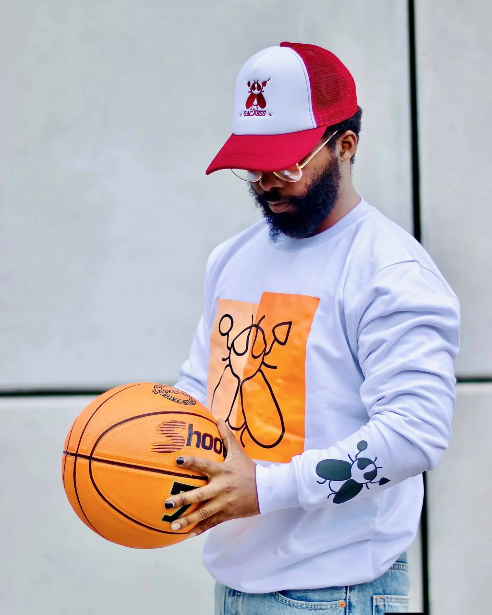 A man in a red and white SACXESS trucker cap and a white beetle-print sweatshirt, looking down at a basketball